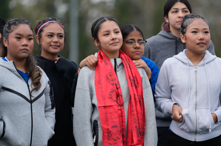 FILE - Samira Abdul rests her head on Valeria Hernandez's shoulder, center, as they stand with teammates before a match at a soccer tournament for immigrant and refugee girls on Sunday, March 29, 2026, in Portland, Ore. Valeria, her mother, and younger sister were gifted brightly colored scarves as symbols of support.Jenny Kane / AP